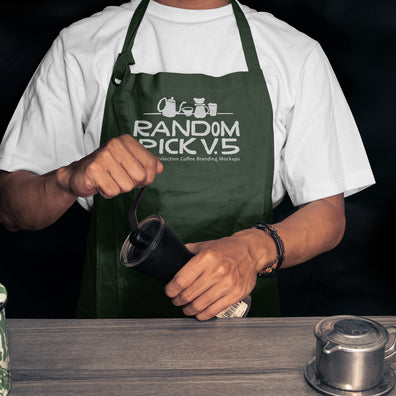 Realistic dark green barista apron mockup worn by a man holding a coffee grinder on a moody dark background with coffee props on a wooden table