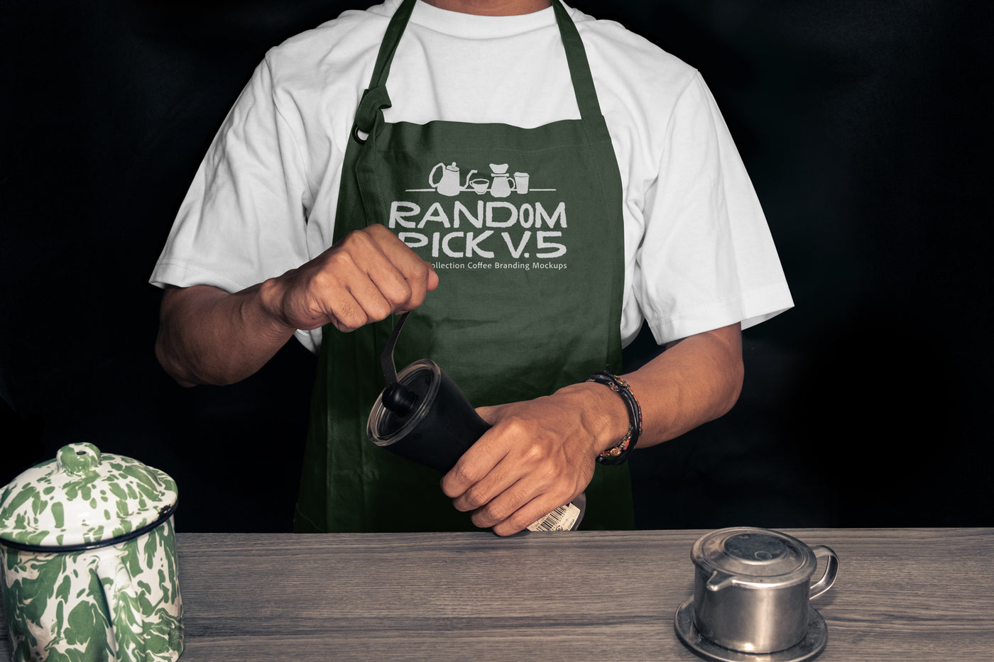 Realistic dark green barista apron mockup worn by a man holding a coffee grinder on a moody dark background with coffee props on a wooden table