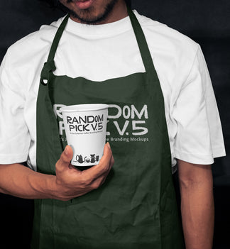 Barista wearing a dark green apron holding a branded paper coffee cup in a moody dark cafe setting, showing both apron and cup branding mockup