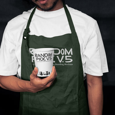 Barista wearing a dark green apron holding a branded paper coffee cup in a moody dark cafe setting, showing both apron and cup branding mockup