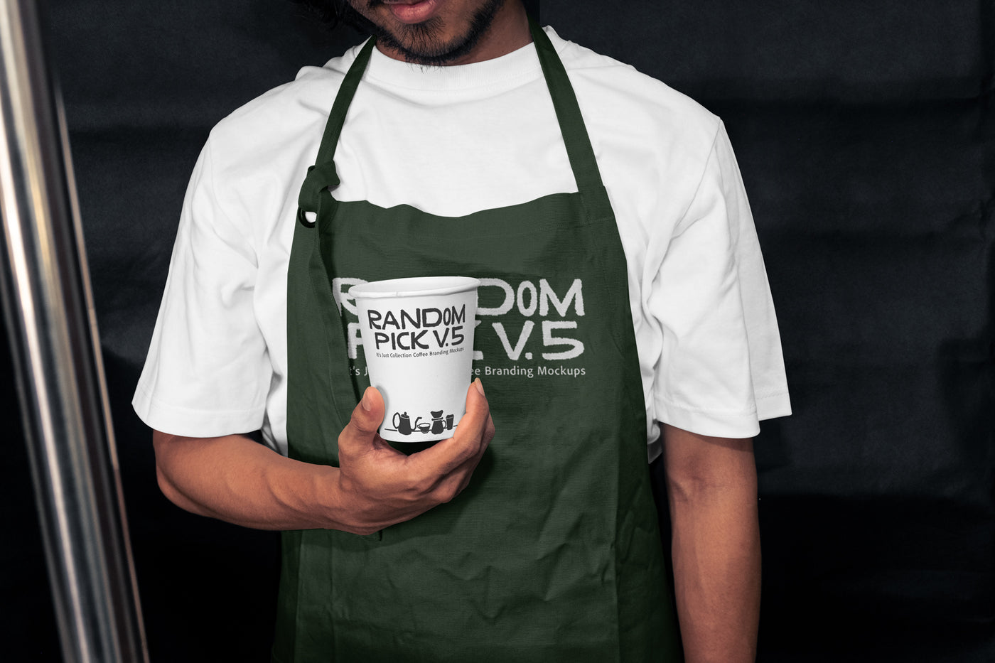 Barista wearing a dark green apron holding a branded paper coffee cup in a moody dark cafe setting, showing both apron and cup branding mockup