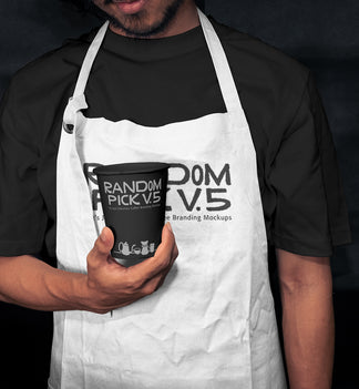 Barista wearing a white apron holding a branded paper coffee cup in a moody dark cafe setting, showing both apron and cup branding mockup