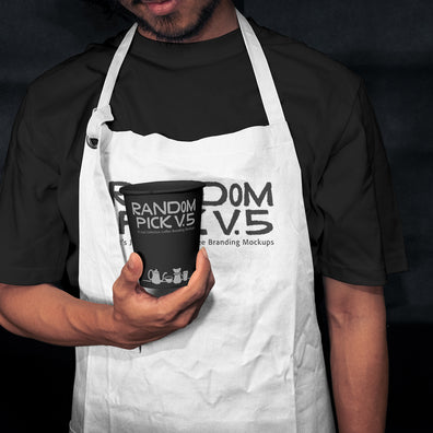 Barista wearing a white apron holding a branded paper coffee cup in a moody dark cafe setting, showing both apron and cup branding mockup