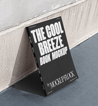 A black book with white text reading THE COOL BREEZE BOOK MOCKUP BY MOCKUP FLOCK is lying on a clean floor, against the wall.