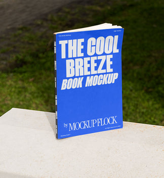 A blue book with white text reading THE COOL BREEZE BOOK MOCKUP BY MOCKUP FLOCK is lying on a fence, displayed in a natural outdoor setting.