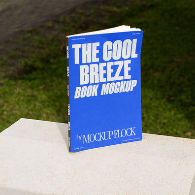 A blue book with white text reading THE COOL BREEZE BOOK MOCKUP BY MOCKUP FLOCK is lying on a fence, displayed in a natural outdoor setting.