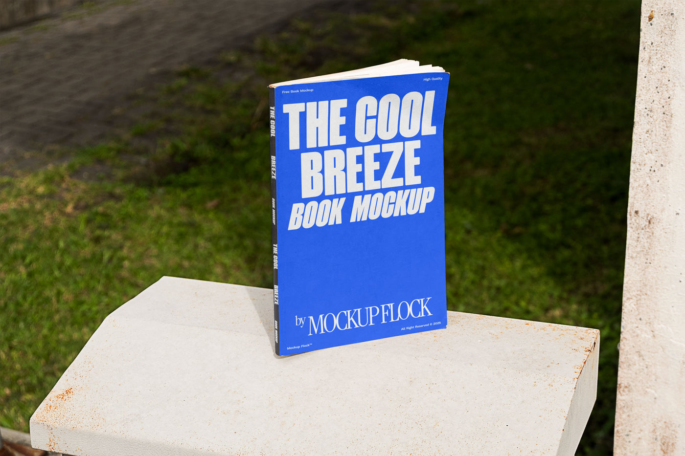 A blue book with white text reading THE COOL BREEZE BOOK MOCKUP BY MOCKUP FLOCK is lying on a fence, displayed in a natural outdoor setting.