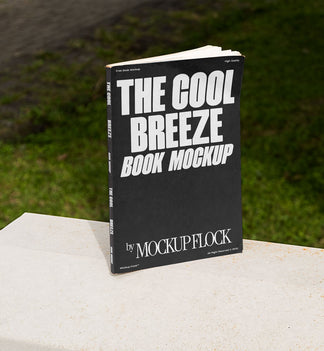 A black book with white text reading THE COOL BREEZE BOOK MOCKUP BY MOCKUP FLOCK is lying on a fence, displayed in a natural outdoor setting.