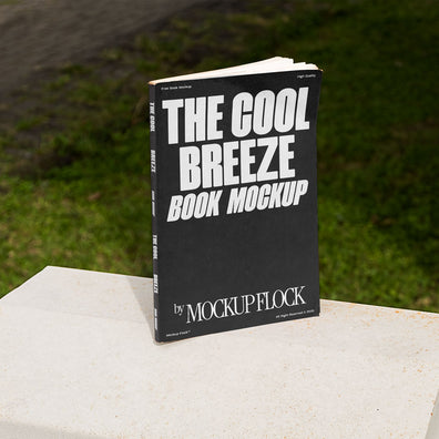 A black book with white text reading THE COOL BREEZE BOOK MOCKUP BY MOCKUP FLOCK is lying on a fence, displayed in a natural outdoor setting.