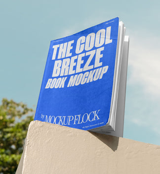 A blue book with white text reading THE COOL BREEZE BOOK MOCKUP BY MOCKUP FLOCK is lying on a fence, displayed in a natural outdoor setting.