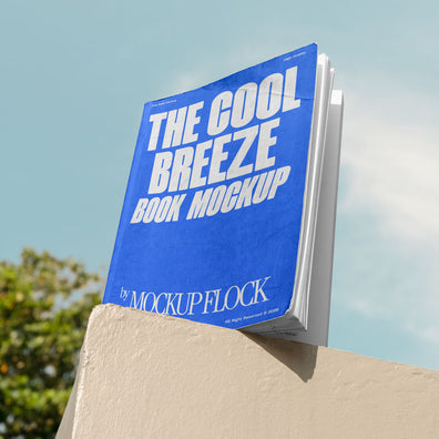 A blue book with white text reading THE COOL BREEZE BOOK MOCKUP BY MOCKUP FLOCK is lying on a fence, displayed in a natural outdoor setting.
