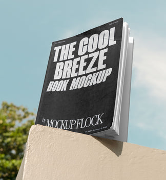 A black book with white text reading THE COOL BREEZE BOOK MOCKUP BY MOCKUP FLOCK is lying on a fence, displayed in a natural outdoor setting.