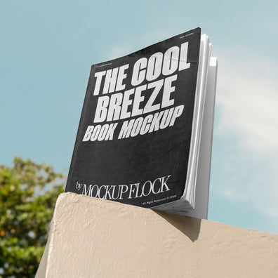 A black book with white text reading THE COOL BREEZE BOOK MOCKUP BY MOCKUP FLOCK is lying on a fence, displayed in a natural outdoor setting.