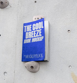 A blue book with white text reading THE COOL BREEZE BOOK MOCKUP BY MOCKUP FLOCK is lying on a rock climbing wall. Design by Mockup Flock