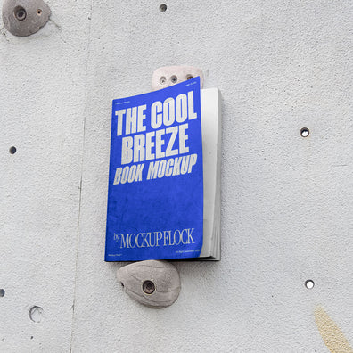 A blue book with white text reading THE COOL BREEZE BOOK MOCKUP BY MOCKUP FLOCK is lying on a rock climbing wall. Design by Mockup Flock