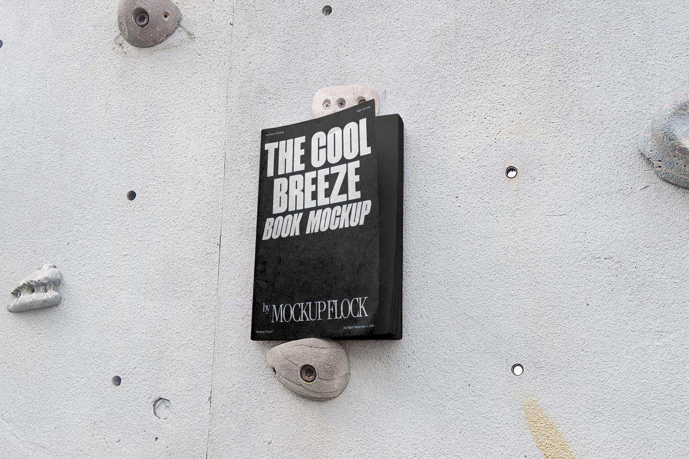 A black book with white text reading THE COOL BREEZE BOOK MOCKUP BY MOCKUP FLOCK is lying on a rock climbing wall. Design by Mockup Flock