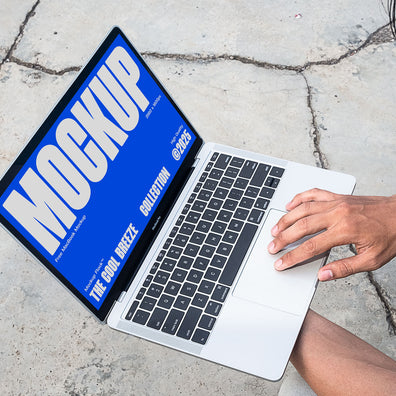A person is holding an Macbook Device that says "MOCKUP THE COOL BREEZE COLLECTION @2025" in bold white text. Design by Mockup Flock