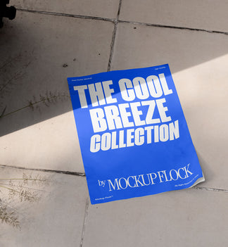 A blue Poster with white text reading THE COOL BREEZE COLLECTION BY MOCKUP FLOCK is lying on a floor, displayed in a natural outdoor setting.