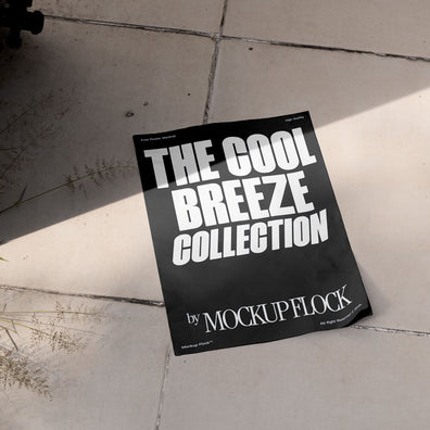 A black Poster with white text reading THE COOL BREEZE COLLECTION BY MOCKUP FLOCK is lying on a floor, displayed in a natural outdoor setting.