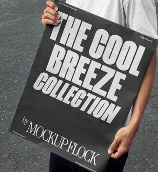 A person holds a poster that reads "THE COOL BREEZE COLLECTION BY MOCKUP FLOCK" in bold text, standing on black asphalt.
