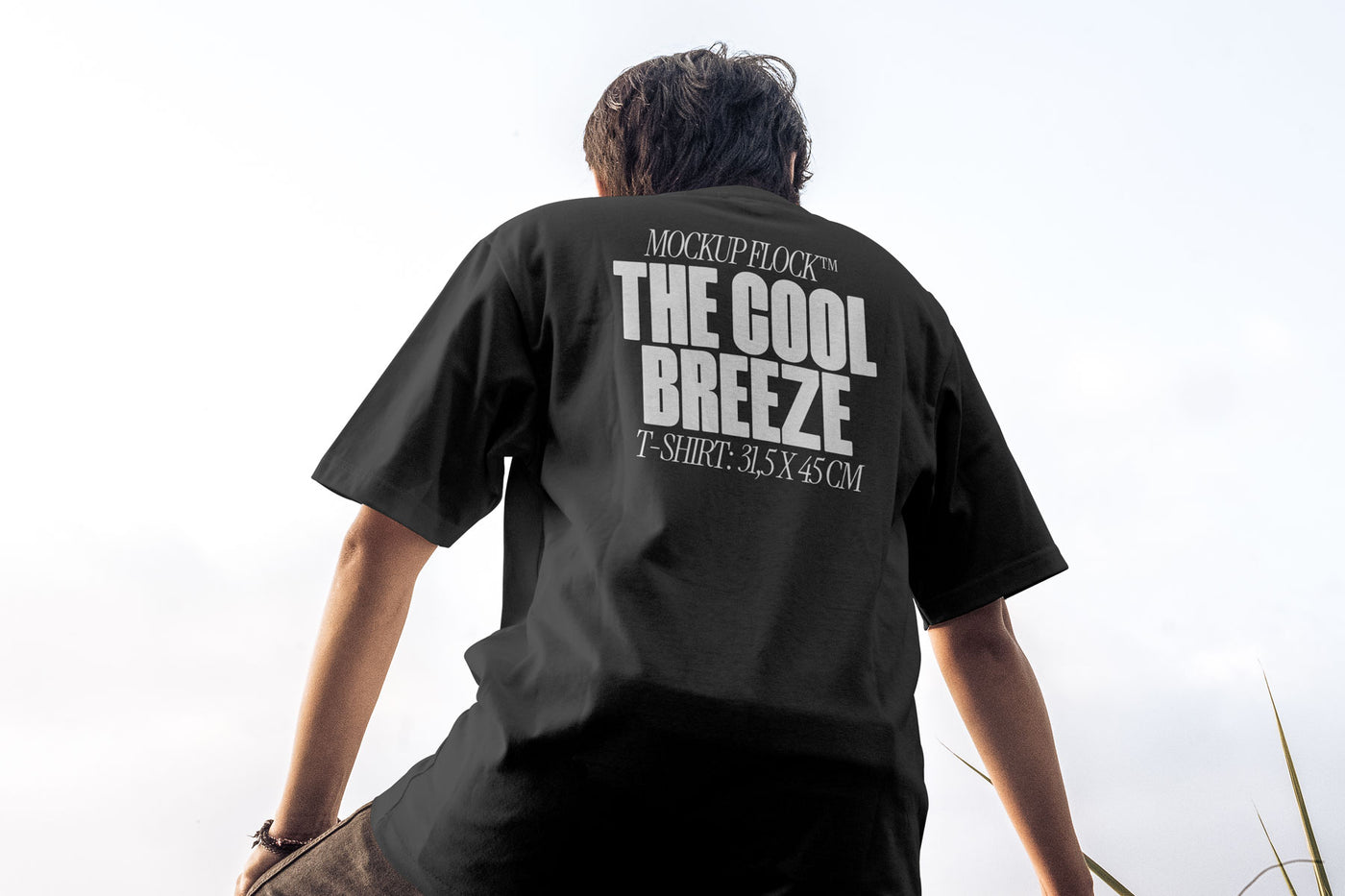 A person wearing a t-shirt that says "MOCKUP FLOCK THE COOL BREEZE T-SHIRT: 31,5X45CM" in bold text, on a blue sky background.
