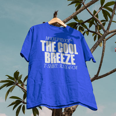 A blue t-shirt with white text that reads "MOCKUP FLOCK THE COOL BREEZE T-SHIRT: 31.5X 45 CM" hanging on a tree, displayed outdoors.