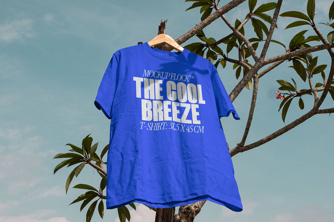 A blue t-shirt with white text that reads "MOCKUP FLOCK THE COOL BREEZE T-SHIRT: 31.5X 45 CM" hanging on a tree, displayed outdoors.