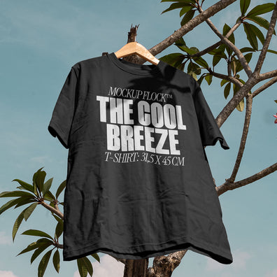 A black t-shirt with white text that reads "MOCKUP FLOCK THE COOL BREEZE T-SHIRT: 31.5X 45 CM" hanging on a tree, displayed outdoors.