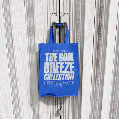 A blue tote bag with white text that reads "THE COOL BREEZE COLLECTION FREE TOTE BAG MOCKUP" hanging on a door handle.