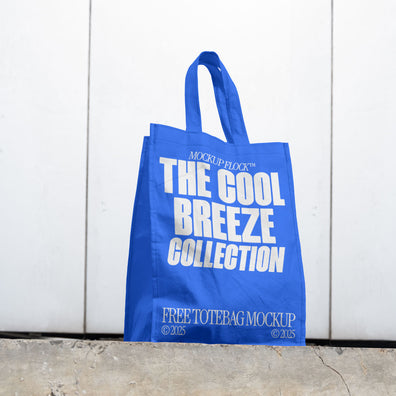 A blue tote bag with white text reading THE COOL BREEZE COLLECTION BY MOCKUP FLOCK is lying on the floor, on a white wall background.