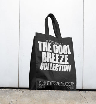 A black tote bag with white text reading THE COOL BREEZE COLLECTION BY MOCKUP FLOCK is lying on the floor, on a white wall background.