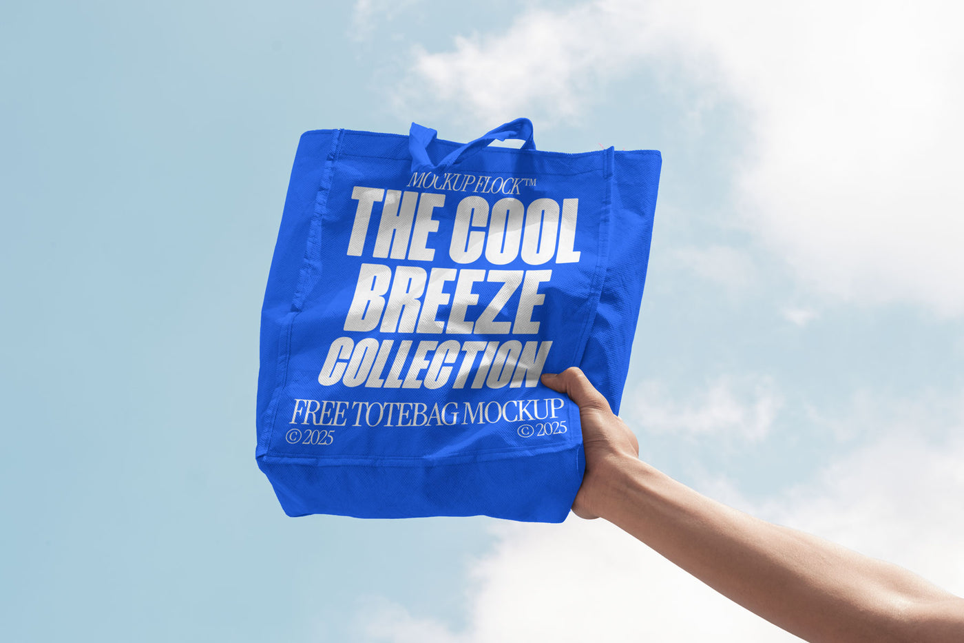 A person holds a tote bag that says "MOCKUP FLOCK THE COOL BREEZE COLLECTION FREE TOTE BAG MOCKUP" in bold text, on a blue sky background.