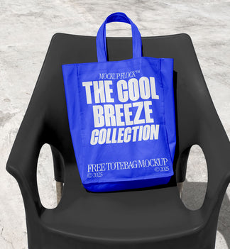 A blue tote bag with white text reading "THE COOL BREEZE COLLECTION FREE TOTE BAG MOCKUP" is lying on black chair.