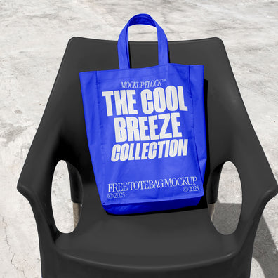 A blue tote bag with white text reading "THE COOL BREEZE COLLECTION FREE TOTE BAG MOCKUP" is lying on black chair.
