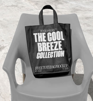A black Poster with white text reading "THE COOL BREEZE COLLECTION FREE TOTE BAG MOCKUP" is lying on grey  chair.