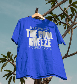 A blue t-shirt with white text that reads "MOCKUP FLOCK THE COOL BREEZE T-SHIRT: 31.5X 45 CM" hanging on a tree, displayed outdoors.