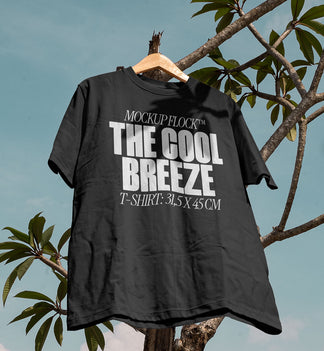 A black t-shirt with white text that reads "MOCKUP FLOCK THE COOL BREEZE T-SHIRT: 31.5X 45 CM" hanging on a tree, displayed outdoors.