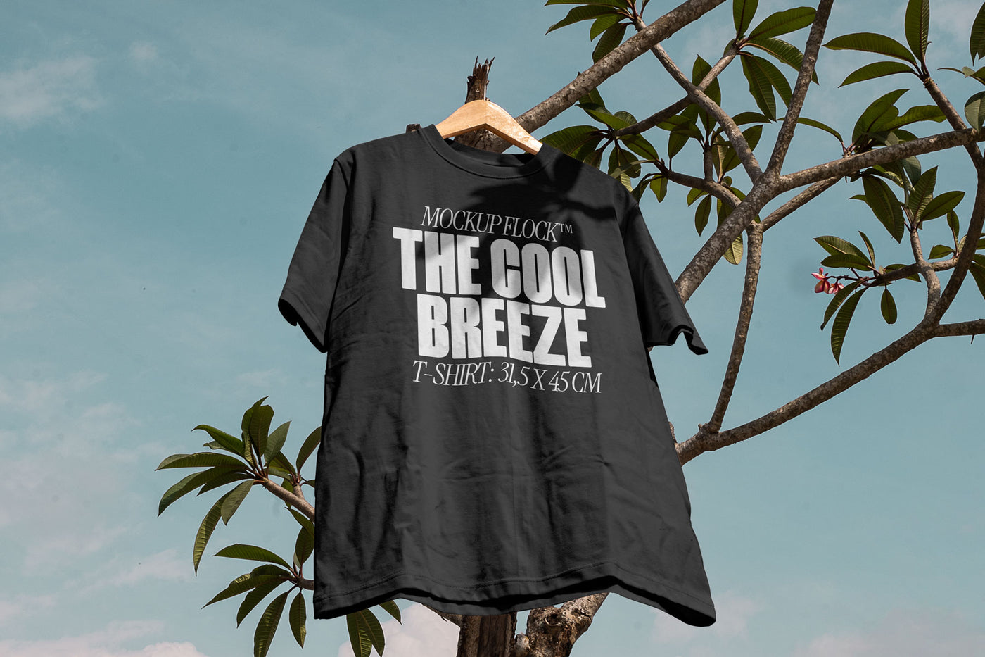 A black t-shirt with white text that reads "MOCKUP FLOCK THE COOL BREEZE T-SHIRT: 31.5X 45 CM" hanging on a tree, displayed outdoors.
