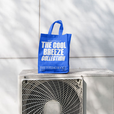 A blue Poster with white text reading "MOCKUP FLOCK THE COOL BREEZE COLLECTION FREE TOTE BAG MOCKUP" is lying on outdoor air conditioner.