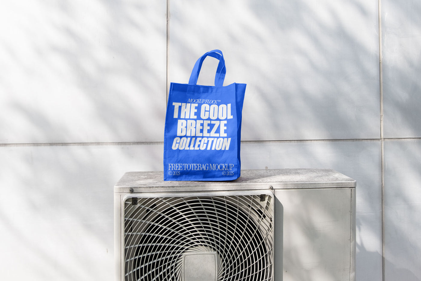 A blue Poster with white text reading "MOCKUP FLOCK THE COOL BREEZE COLLECTION FREE TOTE BAG MOCKUP" is lying on outdoor air conditioner.
