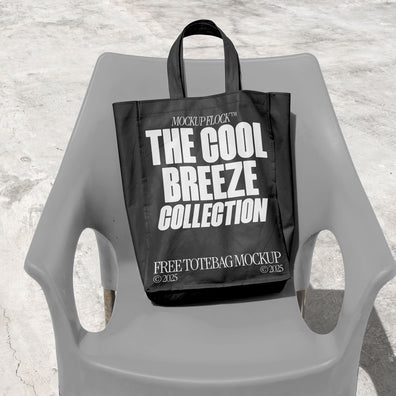 A black Poster with white text reading "THE COOL BREEZE COLLECTION FREE TOTE BAG MOCKUP" is lying on grey  chair.