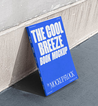 A blue book with white text reading THE COOL BREEZE BOOK MOCKUP BY MOCKUP FLOCK is lying on a clean floor, against the wall.