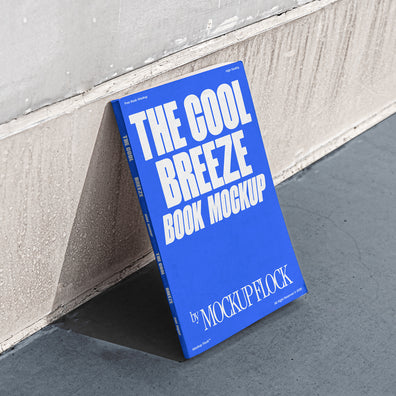 A blue book with white text reading THE COOL BREEZE BOOK MOCKUP BY MOCKUP FLOCK is lying on a clean floor, against the wall.