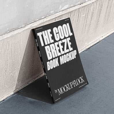 A black book with white text reading THE COOL BREEZE BOOK MOCKUP BY MOCKUP FLOCK is lying on a clean floor, against the wall.