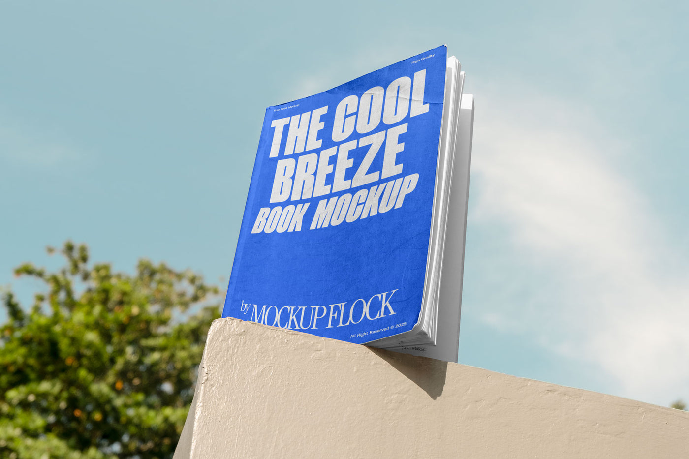 A blue book with white text reading THE COOL BREEZE BOOK MOCKUP BY MOCKUP FLOCK is lying on a fence, displayed in a natural outdoor setting.
