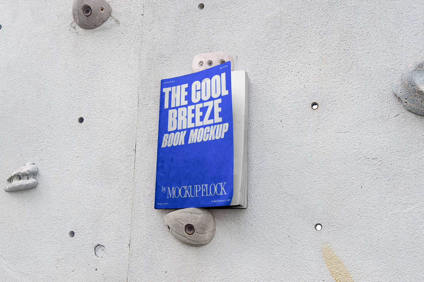 A blue book with white text reading THE COOL BREEZE BOOK MOCKUP BY MOCKUP FLOCK is lying on a rock climbing wall. Design by Mockup Flock
