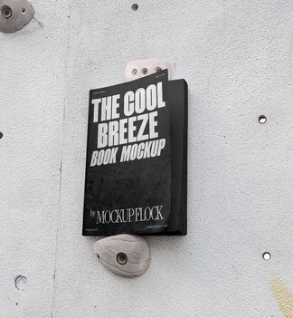 A black book with white text reading THE COOL BREEZE BOOK MOCKUP BY MOCKUP FLOCK is lying on a rock climbing wall. Design by Mockup Flock