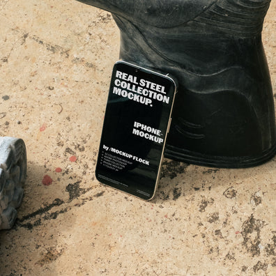 MockupFlock Real Steel Collection iPhone mockup leaning against a black rubber boot on a rough, dusty concrete floor, showcasing realistic lighting and industrial textures.