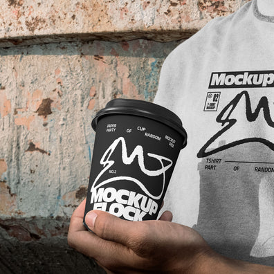 Person wearing a white t-shirt holding a black paper coffee cup with custom branding against an urban textured wall