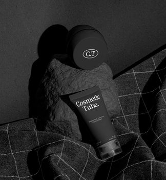 A cosmetic product arrangement featuring a black jar mockup and a black tube mockup labeled 'Cosmetic Tube.' resting on a dark stone against a dark background with draped dark fabric.